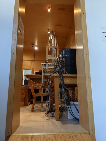 Looking through a door into a 1970s-esque wooden room, featuring a metal-framed tower and wire-sprouting tabletop instrument