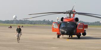 A woman walks towards the camera and away from a large black and orange helicopter on a large landing pad.