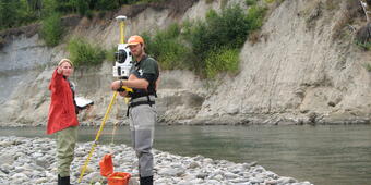 A woman and a man wearing safety gear stand on a rocky embankment along a river near a tripod with a device.
