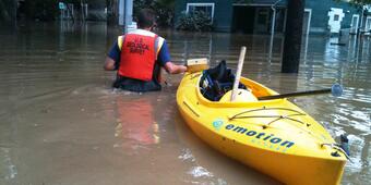 man in orange lifevest in brown floodwaters pulling yellow kayak