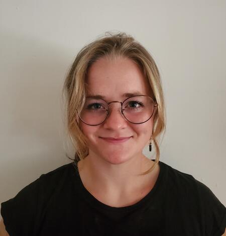 Blonde woman with glasses and black shirt standing against plain background