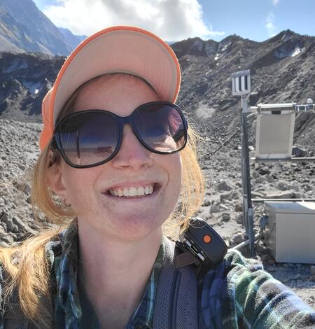 A young woman in dark sunglasses and an orange hat crouches next to a scientific sensor on a rocky slope. She has blonde hair and fair skin.