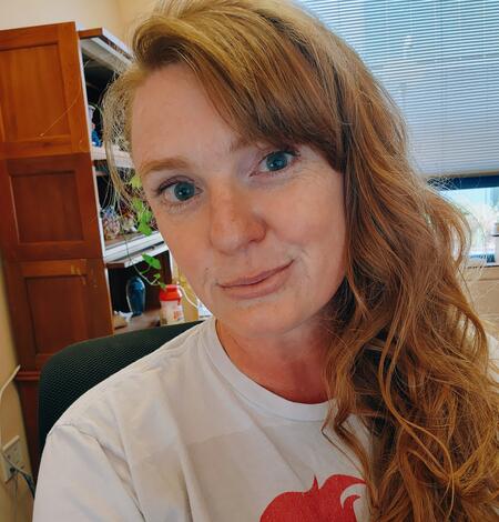 A woman with long hair leans toward the camera with a bookcase and window in the background.