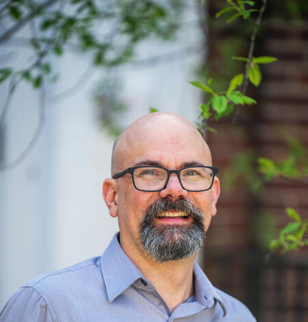Man wearing a blue button-down shirt standing outside with tree behind him