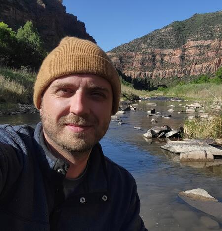 A man wearing a beige cap looks directly at the camera with a stream and red rock canyon behind him