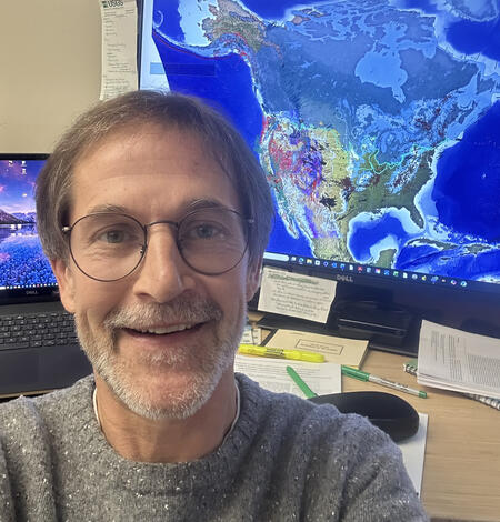 Selfie portrait of Cal Ruleman in front of computer showing geologic map of North America