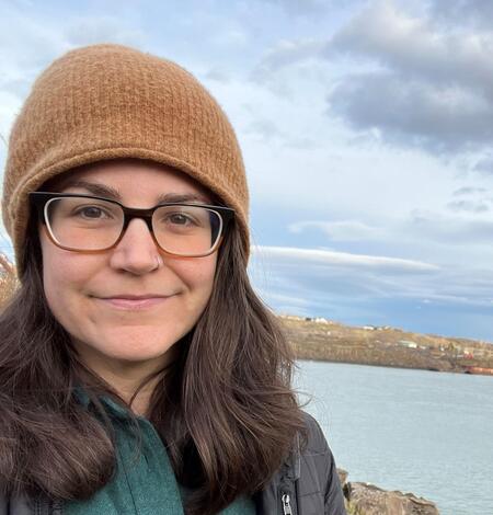 Dark haired female in a tan beanie and glasses stands near the Columbia River on a cloudy day
