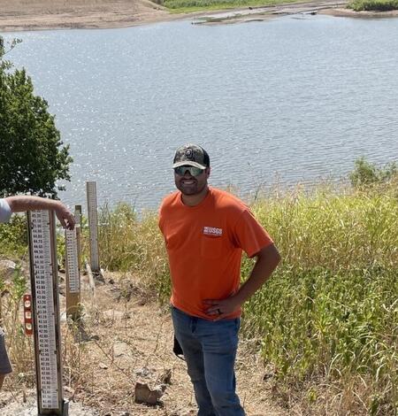 Man with orange USGS shirt, jeans and baseball hat standing at water front