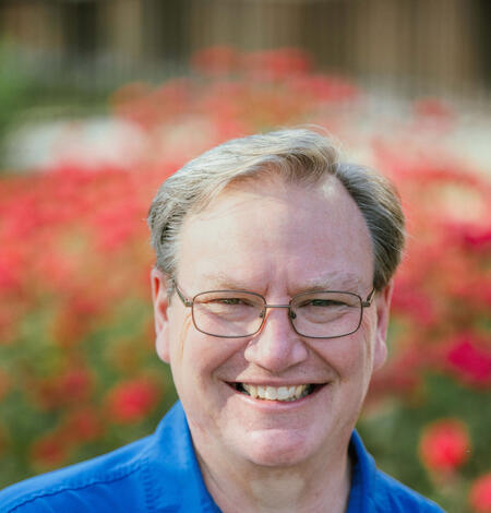 A man in a blue collared shirt wearing glasses and seated in front of red flowers.