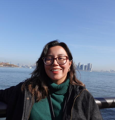 Young woman looking at camera wearing green shirt and black jacket with water in background