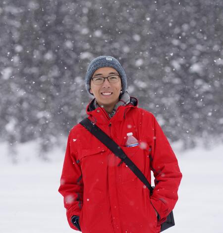 Zehao Xue stands in front of a snowy forest wearing a grey beanie and red jacket.