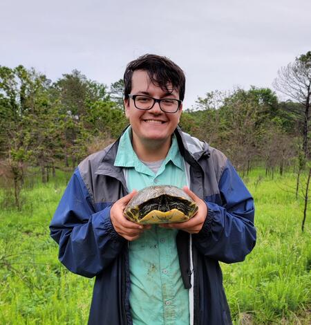 Dylan Wichman photo. Man with dark hair and glasses wearing blue jacket and green shirt standing outdoors holding a turtle