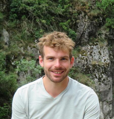 A man in a white t-shirt with curly blonde hair and a beard smiles in front of a vegetation-covered rock face. 