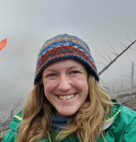 photo of person with long blonde hair in green jacket and multi-colored beanie, tree and clouds behind them