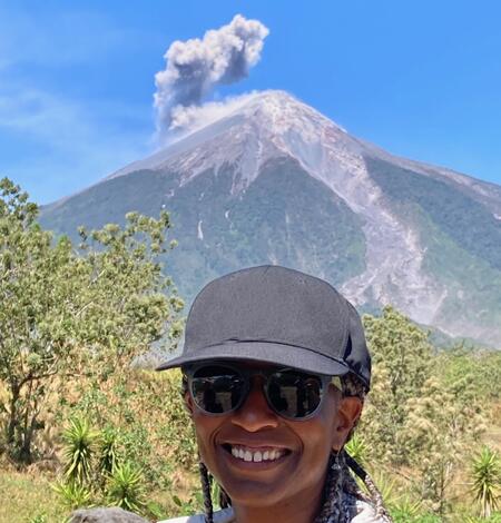 woman with black baseball hat with a volcano erupting ash in the far background.