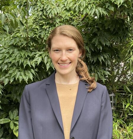 Woman in grey jacket and tan shirt standing in front of green plants.