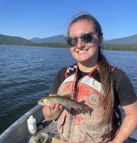 Girl with sunglasses and life jacket holding a fish on a boat. Water, trees, and mountains in the background.