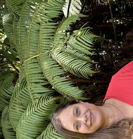 Person in orange shirt with long brown hair in front of tree ferns.
