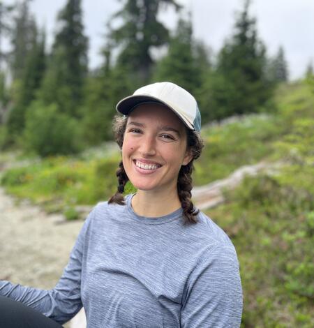 Callie is in a grey long sleeved shirt with a white ball cap and brown hair braided on either side of her face.
