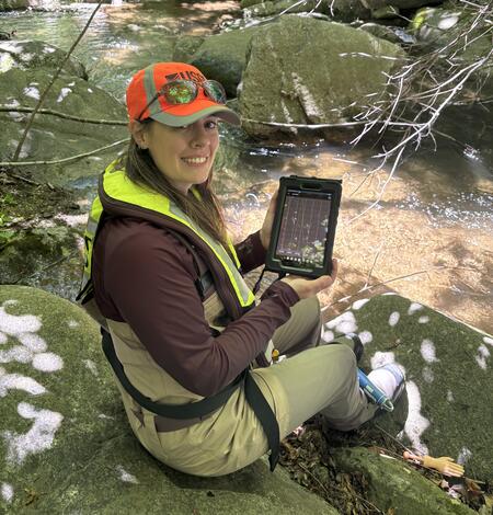 A biologist wearing an orange hat and yellow PFD, sits by a stream holding a tablet.