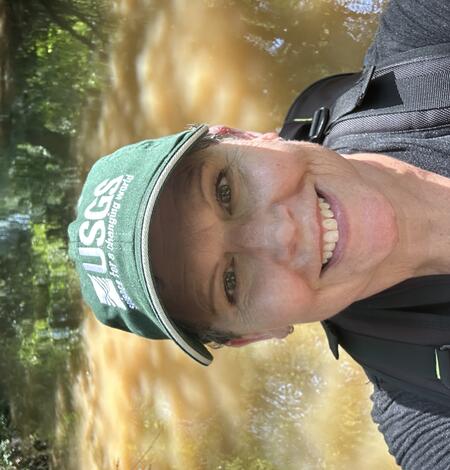 A smiling woman wearing a green USGS baseball hat standing in water and wearing a camera backpack. 