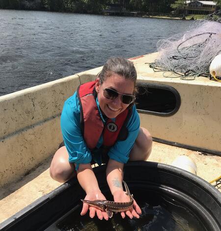 A person in a blue shirt and orange life jacket is on a boat, leaning over a tub of water, holding a small fish in their hands
