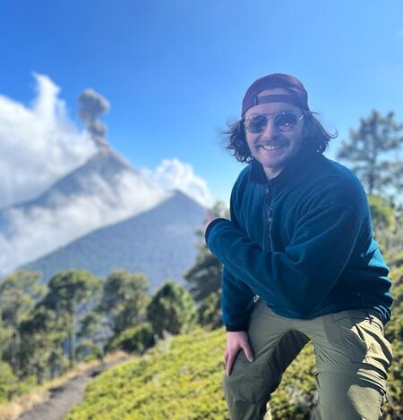 male scientist wearing a backwards hat and sunglasses, pointing towards and erupting volcano in the background.