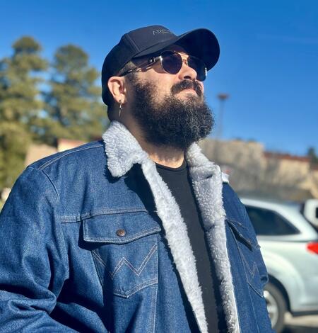 Kelvin, a bearded man with sunglasses, a black cap, and a denim jacket, looking up into the sky