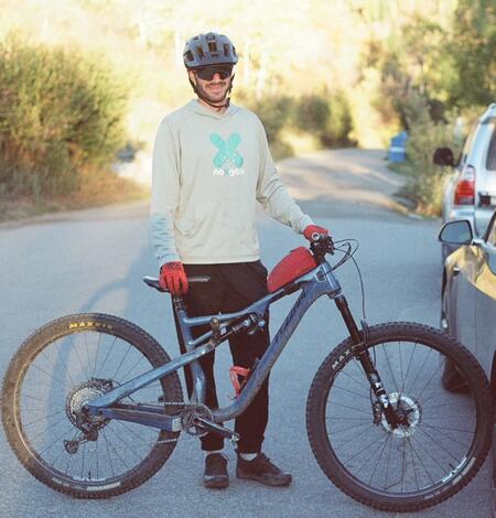 Man wearing bicycle helmet, gloves, and sunglasses stands behind bicycle, trees and car in background