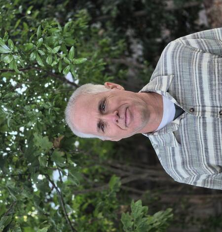Smiling man in button up green and white striped shirt. Green trees form the back ground