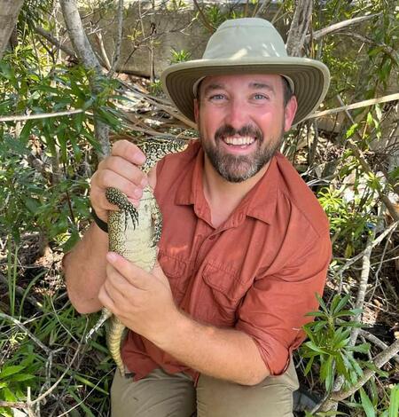 a man in a salmon-colored shirt and a tan hat holds a large black and white lizard, shrubs in the background