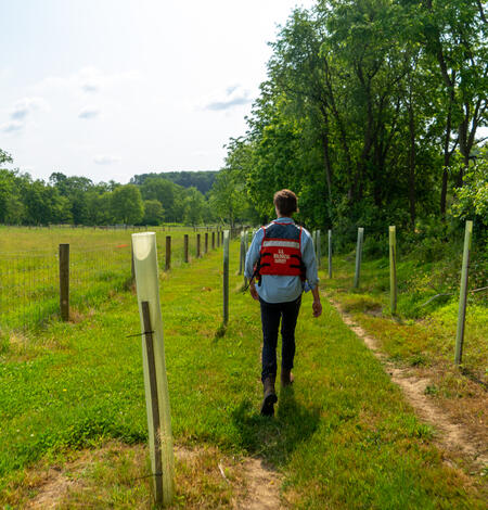 A man wearing an orange life jacket walks down a field with fencing on the left and trees on the right.