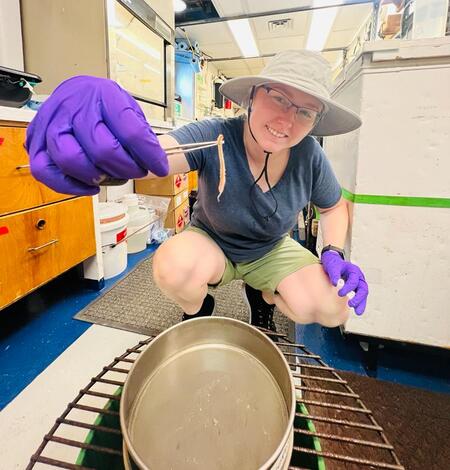 A person in a hat crouches down while holding a worm in their hand and smiles to the camera