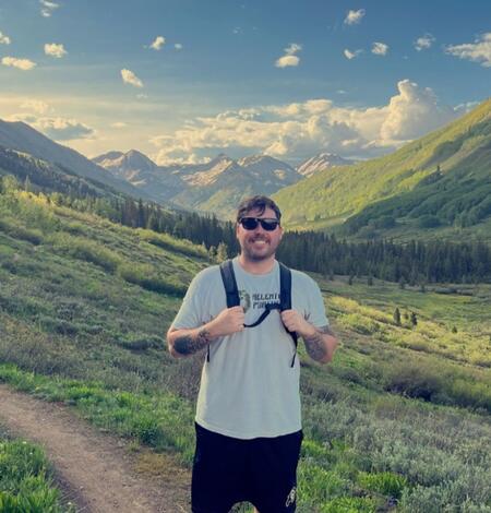Male with brown hair in a grey shirt, black shorts, and glasses smiles amidst stunning mountain scenery