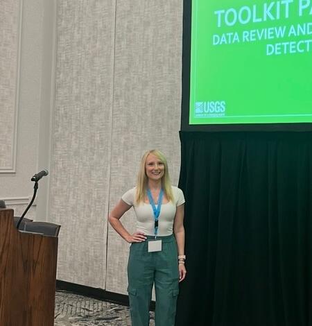 Woman with light skin and long blonde hair standing Infront of a presentation screen and pdium