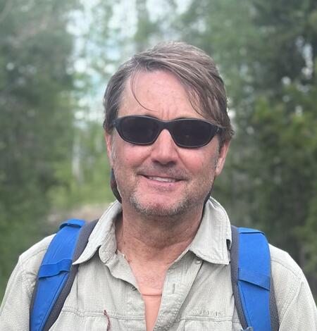 photo of a man's head, man wearing sunglasses and blue backpack straps, trees in the background