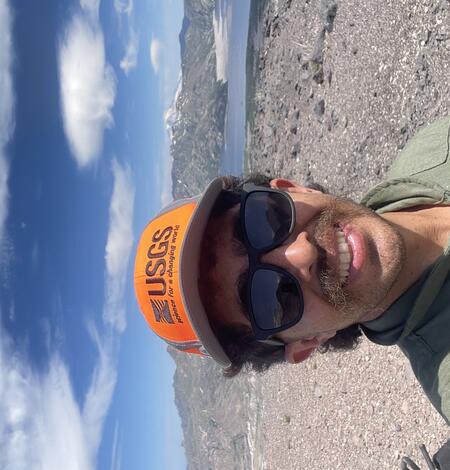 A young man in a green flight suit, orange hat, and dark sunglasses stands on a rocky mountain near a lake. He has brown hair and mustache and fair skin.