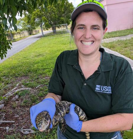 a woman in a dark green shirt and purple gloves holds a black and white lizard, pink building, grass, and trees behind