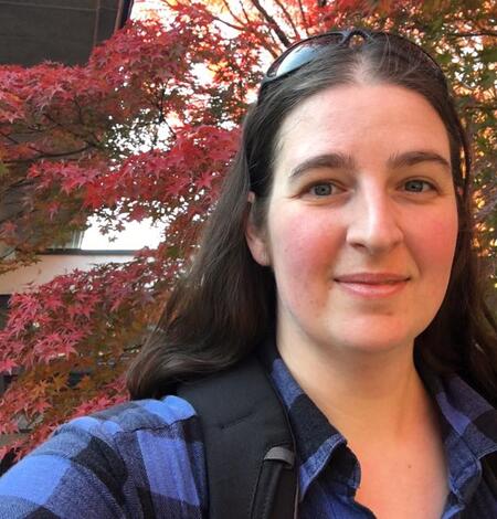 Woman with long brown hair and sunglasses, wearing a flannel shirt. 