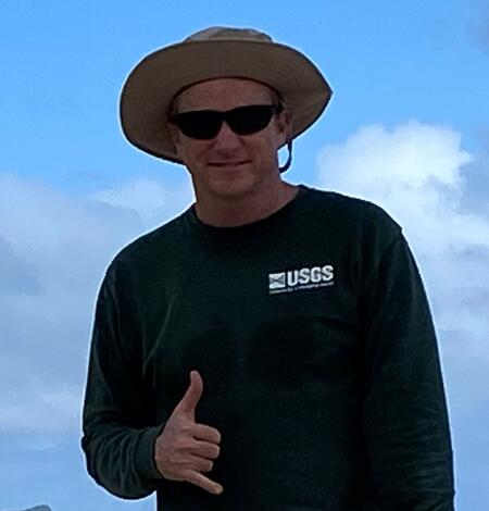 man in long sleeve green USGS shirt and wide brimmed tan hat and sun glasses give the hang loose sign. 