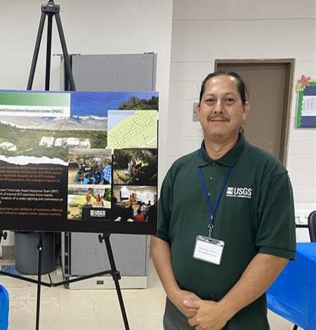 Two people in green shirts stand on either side of a poster inside a community center room.