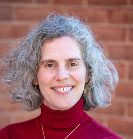 Headshot of a woman with chin-length curly grey hair wearing a maroon turtleneck in front of a brick wall.