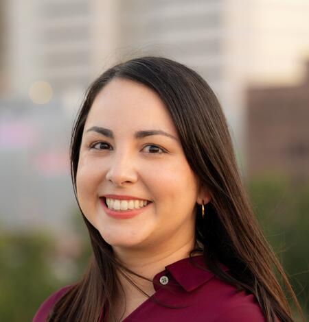 Color photo of Zulimar smiling. She has long brown straight hair and is wearing a marroon shirt with city buildings in the background. 