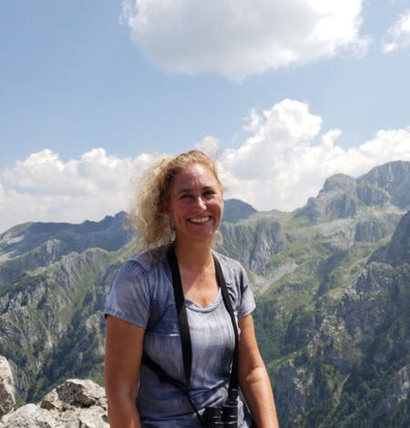 A woman with blonde hair smiles towards the camera with mountains behind her