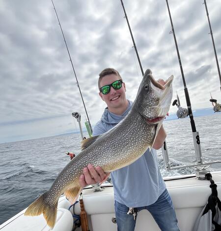 A person on a boat holds up a large fish with speckled coloring and a golden tail. Several fishing rods are positioned upright at the back of the boat, and open water extends into the distance beneath a cloudy sky. 