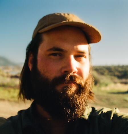 headshot of a male with dark brown beard and tan hat. Outdoors on a sunny day.