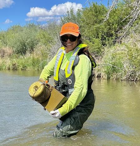 A person wearing waders, PFD, and a USGS hat is standing knee-deep in a river holding a passive pesticides sampler.