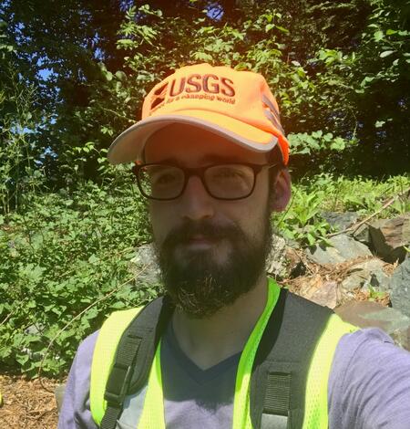 Photo of Alex Bissell standing in the field. The sun is in his eyes and he is smiling. High vis gear is everywhere. 