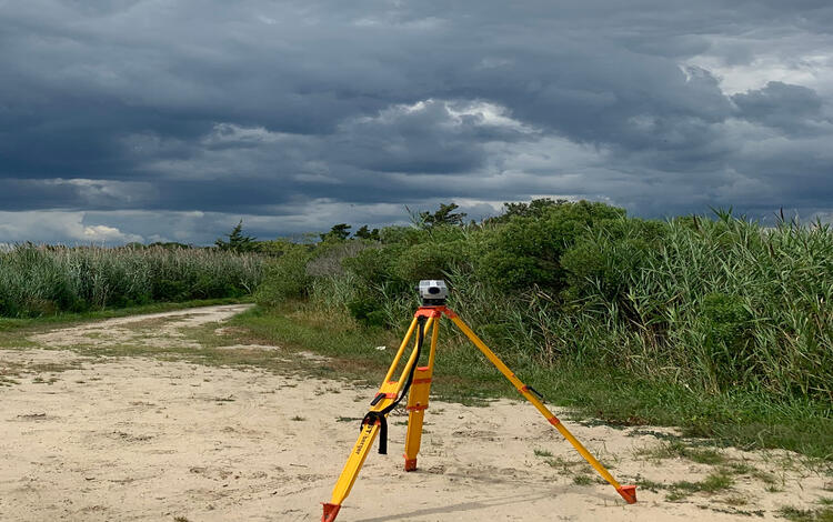 bright yellow tripod with a level transit on top sitting on a sandy beach with high grasses and grey clouds in the background
