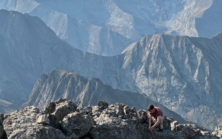 A person is crouched on the ground atop a rocky mountain ridge with numerous ridges seen off in the distance. 
